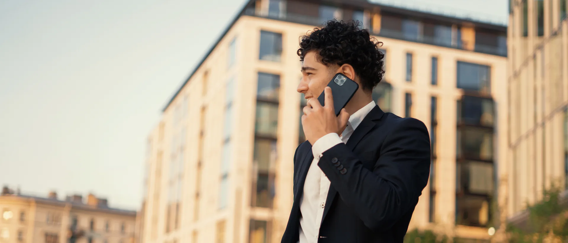 Un uomo giovane in abito blu scuro e camicia bianca cammina per una piazza di giorno con il telefono all’orecchio, di profilo, con un’espressione distesa